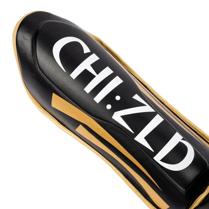 Close-up of shin panel highlighting raised central ridge, CHIZLD word-mark and matte-gold slash graphics on black surface.