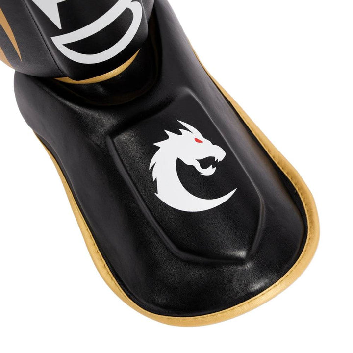 Instep pad close-up: molded foot protector with white dragon-head logo and gold trim framing black synthetic leather.