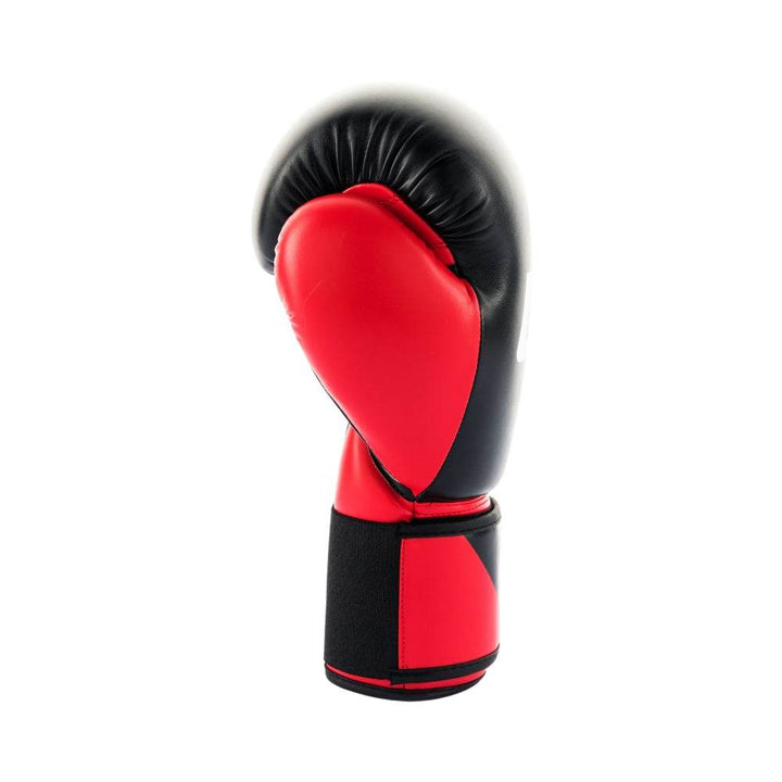 Side view of the red-and-black UFC Pro Compact bag glove’s thumb and wrist area, showing the contoured red thumb,  bold red padding, tight contrast stitching, and the edge of the red hook-and-loop strap folded back for a clear view of the glossy black knuckle panel.