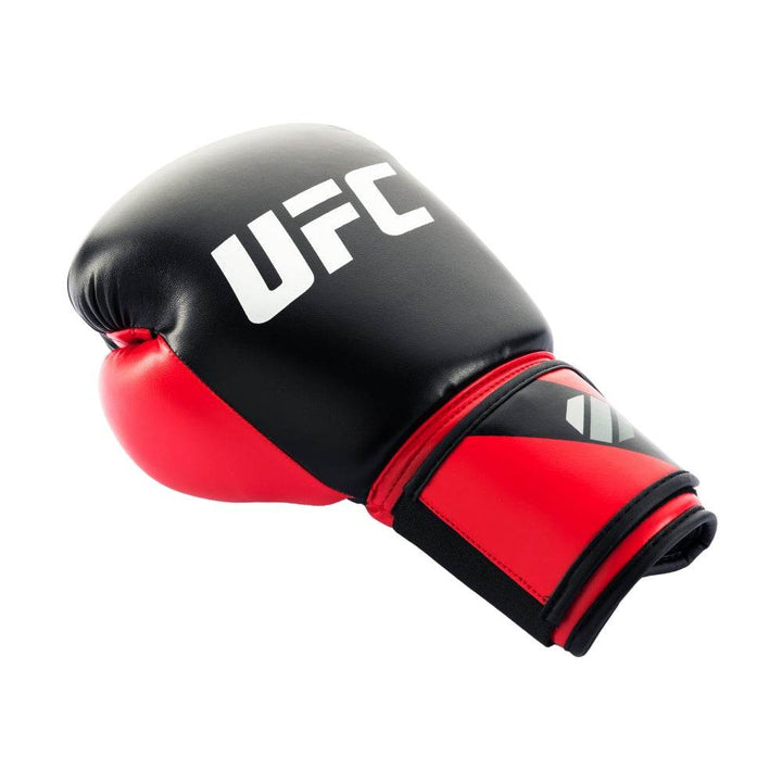 Angled side profile of a red-and-black UFC Pro Compact bag glove resting on its edge, showcasing the curved glossy black knuckle panel with bold red “UFC” lettering, red padded wrist cuff, and partially visible ventilated palm mesh.