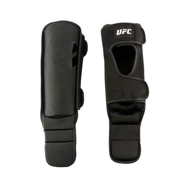 Pair view – front & rear: Two UFC shin guards laid side-by-side. Left guard shows smooth, matte-black strike surface with a faint glossy tonal UFC octagon logo; right guard shows rear side with wide cut-out for the calf, two hook-and-loop straps, and a white “UFC” patch at top.