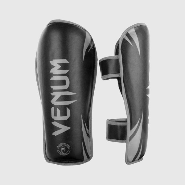 Venum challenger black shin guards with grey detailing shown from front and side angles, featuring bold VENUM branding and dual strap closures.