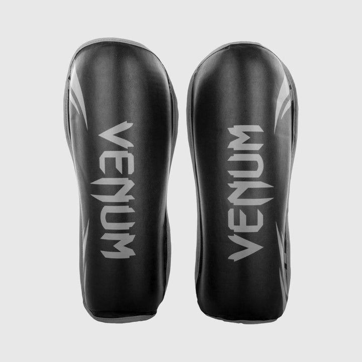 Pair of Venum challenger shin guards in black with grey logo and accents, displayed side-by-side with symmetrical branding.