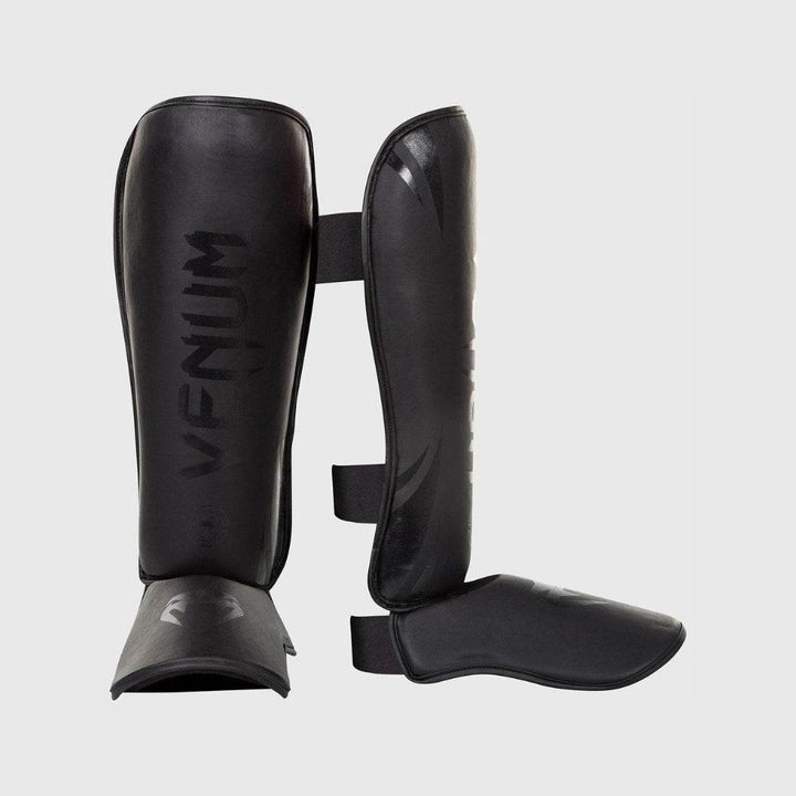 A pair of Venum Challenger Standup Shin Guards shown from front and side angles, featuring matte black finish and subtle gloss logo detailing.