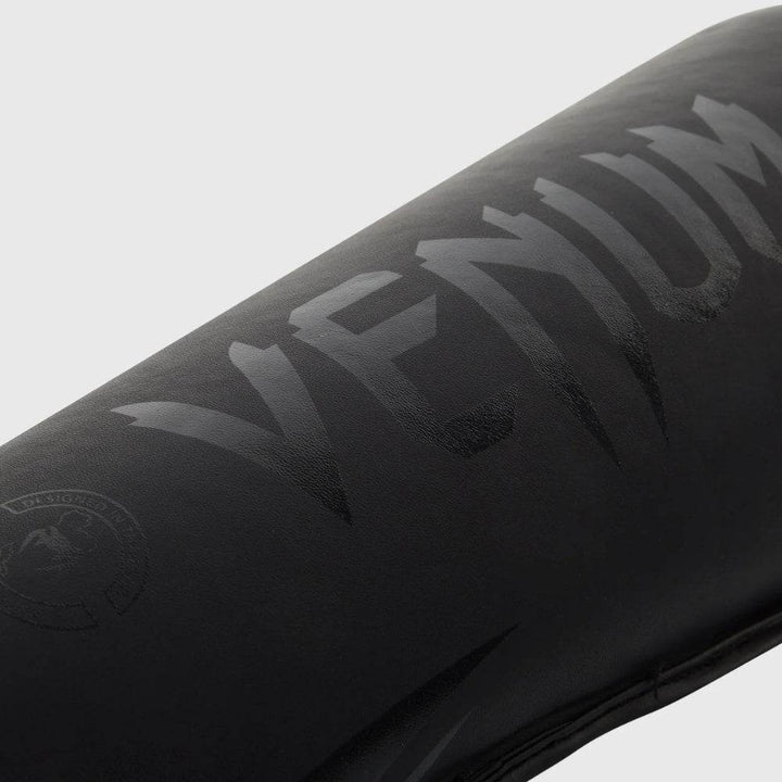 A detailed view of the embossed black-on-black Venum logo printed on the upper shin area of the protective guard.