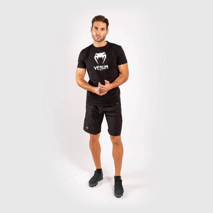 Full body view of the man in black Venum t-shirt and black shorts standing with hands clasped.
