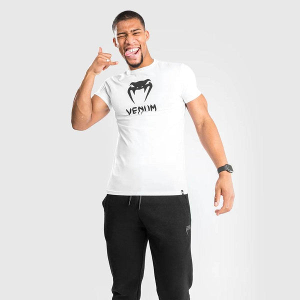 Man in white Venum t-shirt and black joggers making a playful hand gesture while smiling.
