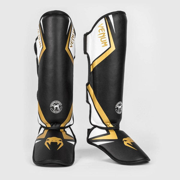 Black, white, and gold Venum Contender 2.0 standup shinguards shown from the front and side angles, highlighting the ergonomic curve and bold snake fang logo on the foot padding.