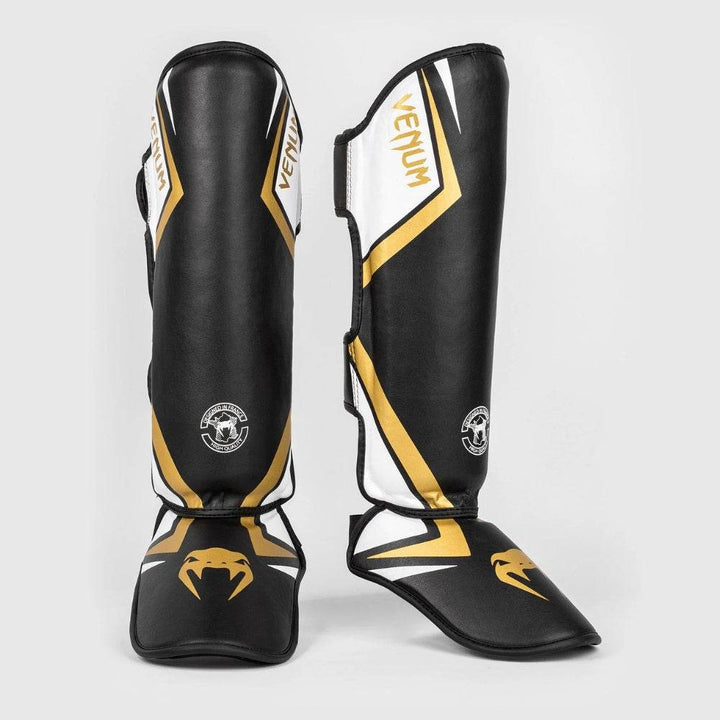 Black, white, and gold Venum Contender 2.0 standup shinguards shown from the front and side angles, highlighting the ergonomic curve and bold snake fang logo on the foot padding.