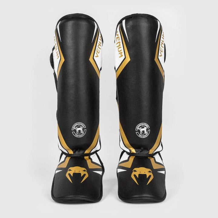 Pair of Venum Contender 2.0 shinguards in black with white and gold accents, displayed front-on to showcase the symmetrical branding and aggressive styling.