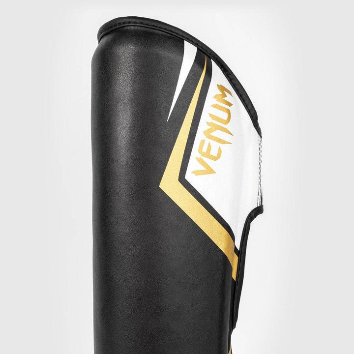 Close-up side view of the upper shinguard area featuring angled white and gold panels with the Venum logo printed in gold.