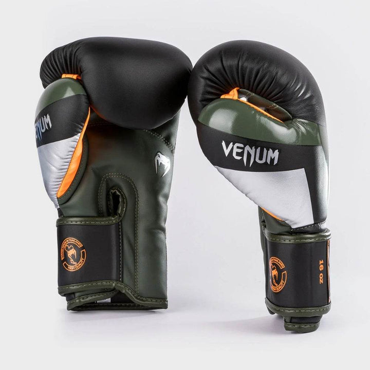 Side profile of Venum Elite gloves in black, silver, and kaki green, showing the thumb and Velcro strap area with orange Venum detailing.