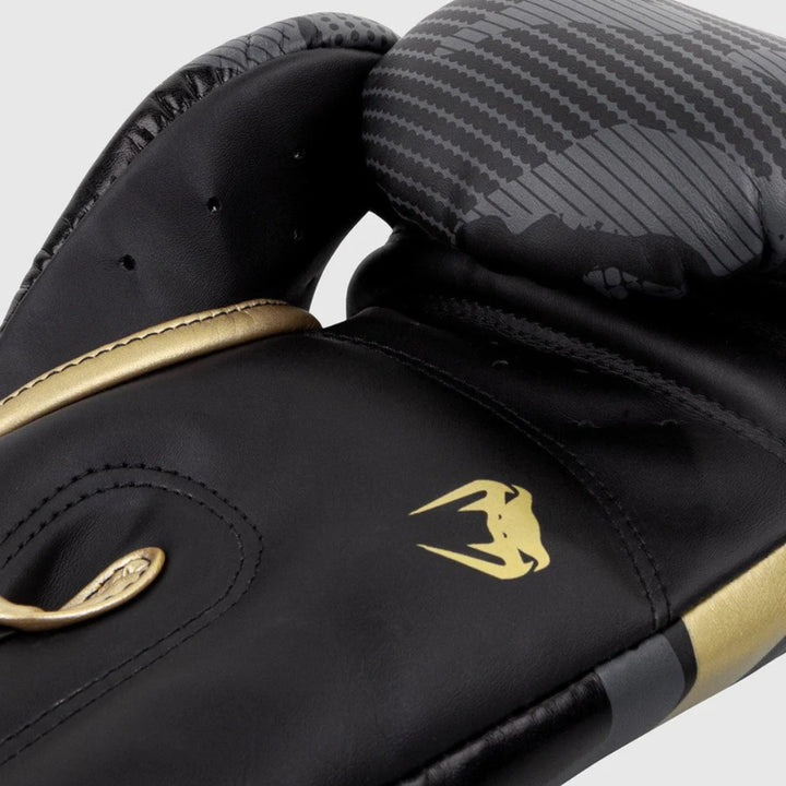 Close-up view of the palm side of a Venum Elite Boxing Glove in Dark Camo/Gold, showing perforations for ventilation, a curved thumb design, and the iconic Venum snake logo on the gold and black surface.