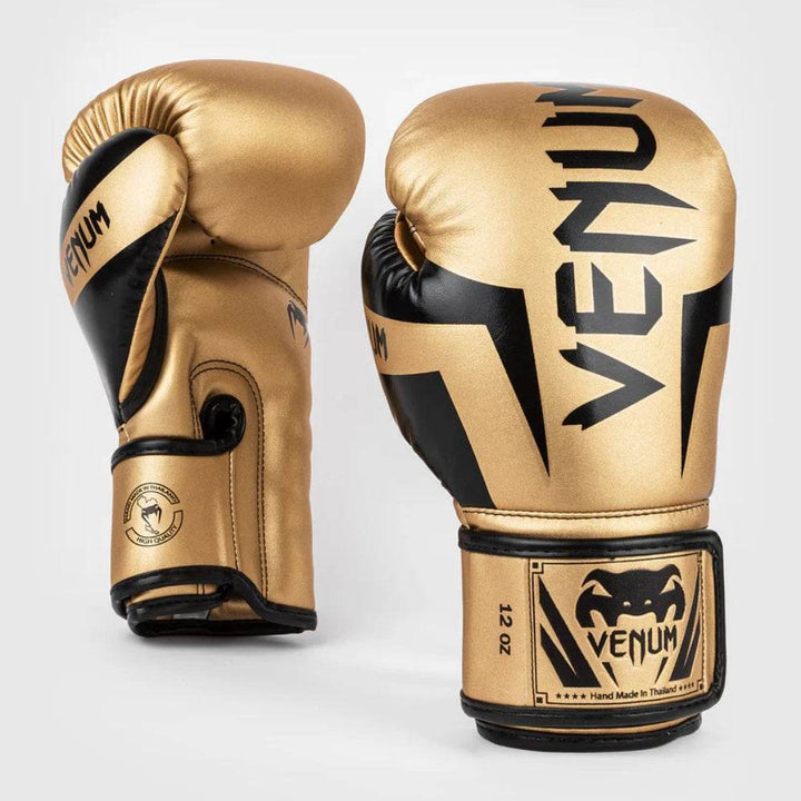 A pair of Venum Elite boxing gloves in a gold and black color scheme, positioned upright. The front glove highlights bold black “VENUM” branding, and the side glove shows the snake logo and wrist design.
