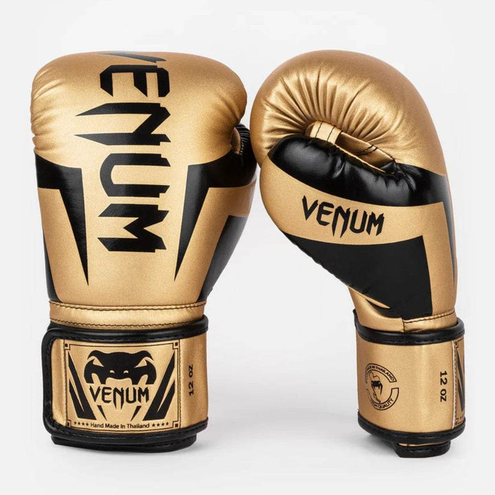 Front and side facing view of Venum Elite boxing gloves in Gold and black. The gloves feature a sharp black pattern on the knuckle area and the signature “VENUM” text vertically and same on the thumb.
