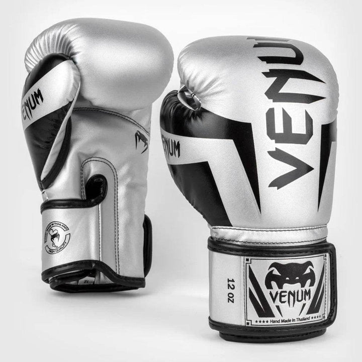 A pair of Venum Elite boxing gloves in a silver and black color scheme, standing upright. The front glove shows bold black “VENUM” branding, while the side glove displays the snake logo and wrist details.
