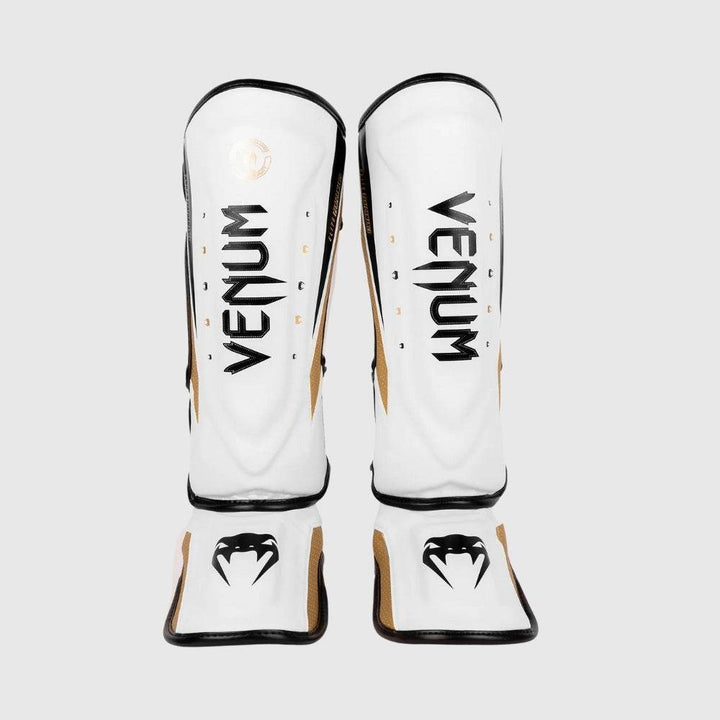 Front view of a pair of white Venum Elite Evo shinguards with bold black Venum logos and gold snakehead graphics on the instep guards.