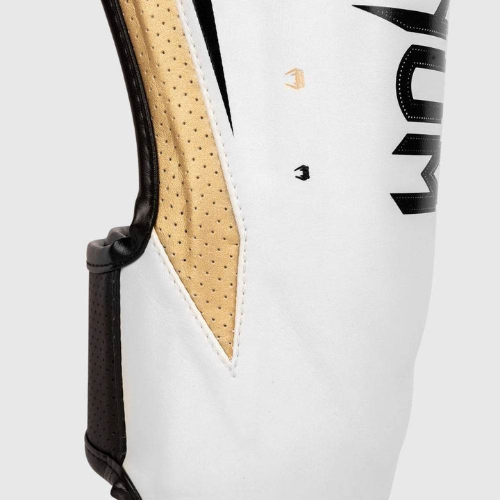 Detailed side view of Venum Elite Evo shinguards showing perforated gold mesh panel and stitching on white leather surface.