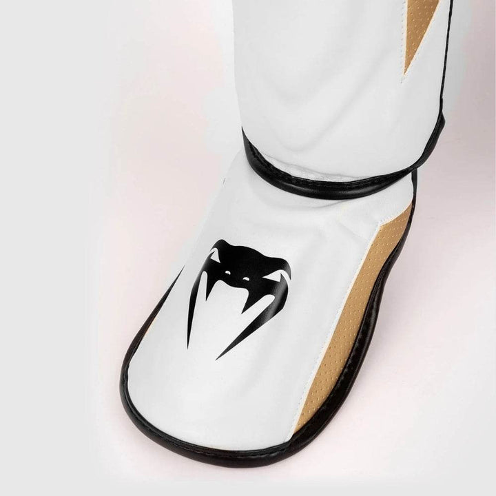 Close-up of the instep section of the Venum Elite Evo shinguard in white with a black Venum snakehead logo and gold mesh sides.
