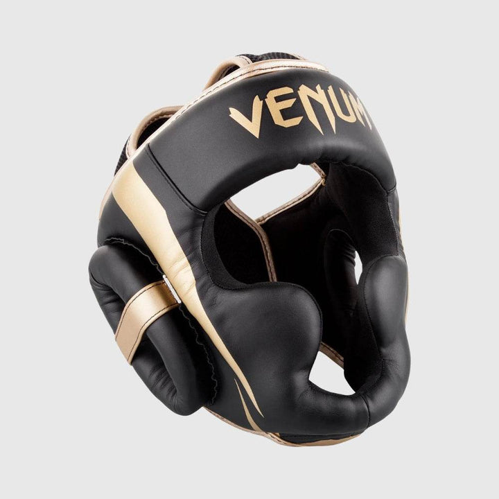Black and gold Venum Elite headgear shown from a front-left angle, highlighting the cheek protection, ear padding, and iconic Venum branding on the forehead.