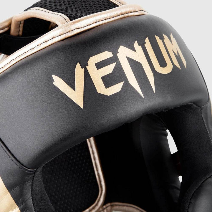 Close-up of the gold “VENUM” logo printed across the forehead padding of the headgear, displayed on a matte black surface.