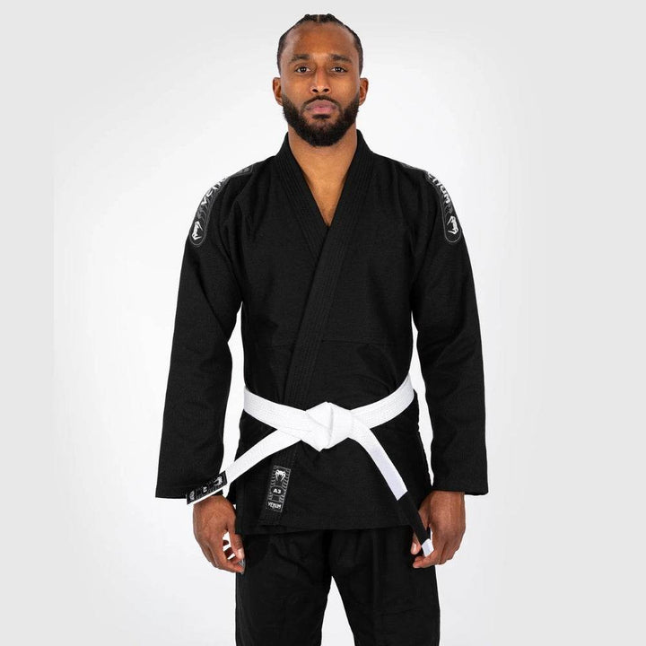 Frontal view of a male model standing straight in the black Venum First BJJ Gi with a white belt, showcasing the clean silhouette and contrast branding.