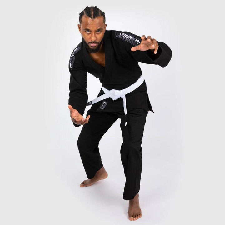 Model in an aggressive grappling stance wearing the black Venum BJJ Gi, demonstrating athletic posture and fit during motion.