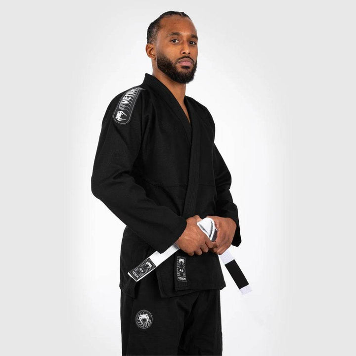 Side profile of the model standing confidently in the black Venum gi, holding the white belt, with a focus on the shoulder patch and overall structure.