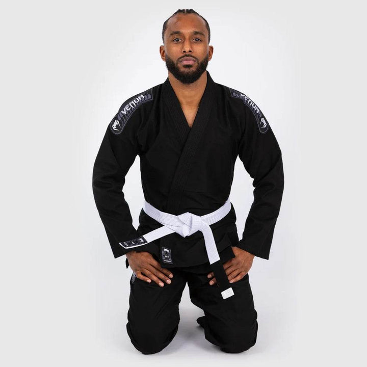 Model kneeling with hands on thighs in the black gi, clearly displaying the front embroidery, belt, and patch placement.