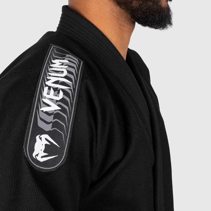 Close-up of the embroidered Venum branding patch on the gi shoulder, emphasizing the white logo over a textured black fabric.