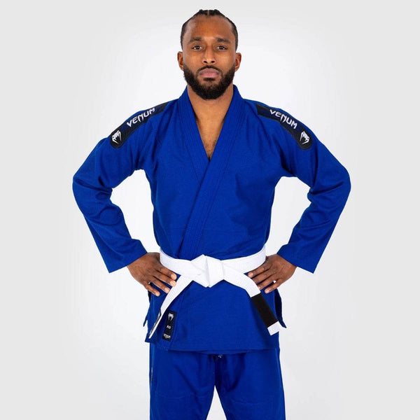 Male model standing upright wearing a blue Venum First BJJ Gi with a white belt, hands on hips, facing forward.