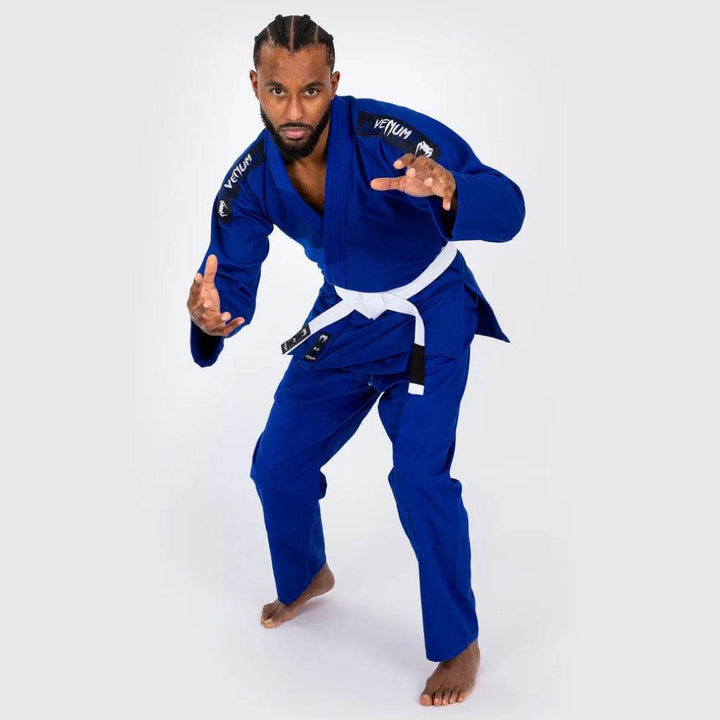 Male model in blue Venum BJJ Gi and white belt in a grappling stance, hands extended forward, ready posture.