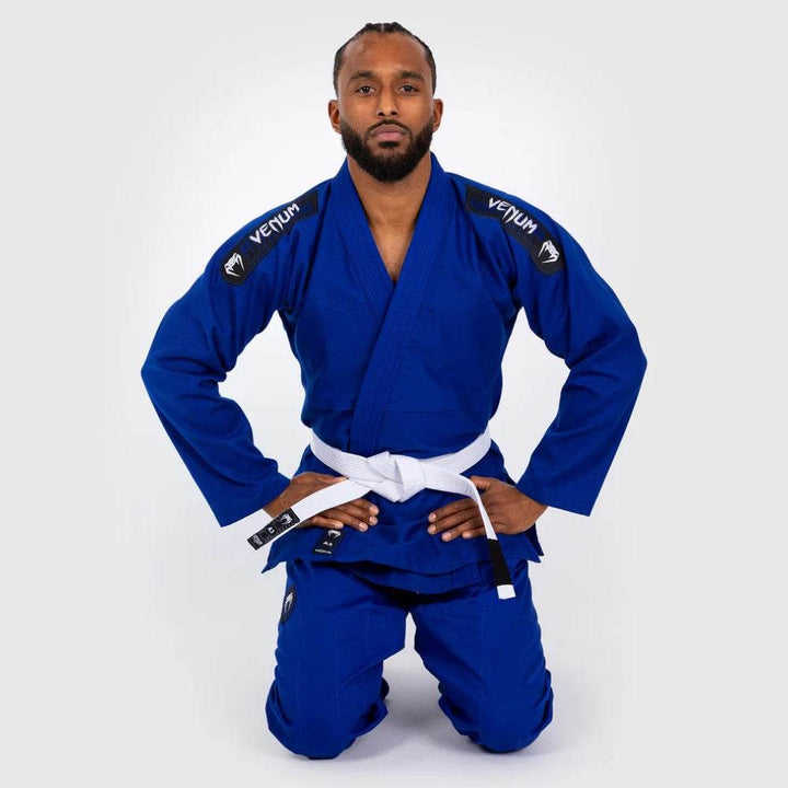 Model kneeling in formal posture wearing blue Venum BJJ Gi and white belt, hands resting on thighs.