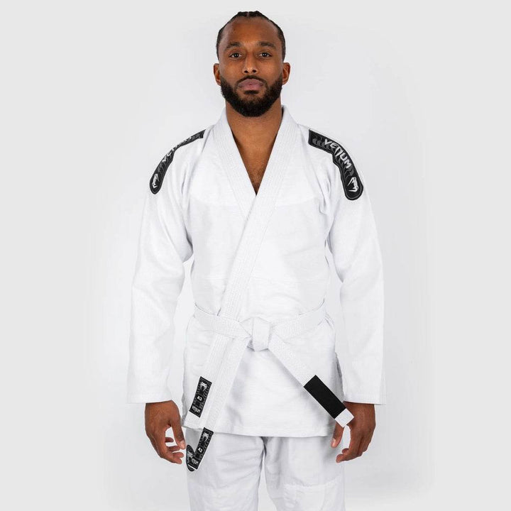 A male martial artist stands upright wearing a white Venum BJJ gi with black shoulder patches and a white belt, facing forward with hands relaxed by his sides.