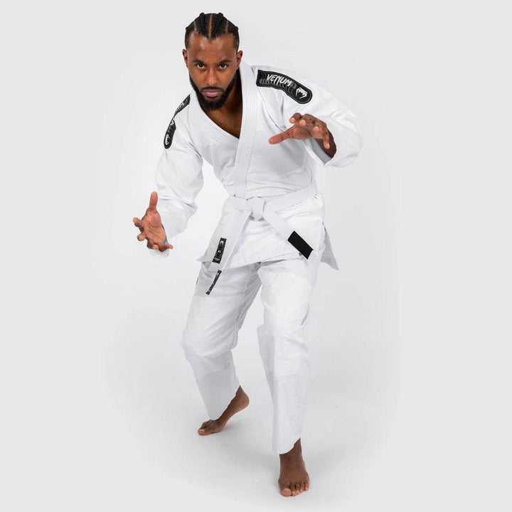 The model poses in a dynamic fighting stance, slightly crouched with arms extended, showcasing the flexibility and fit of the white Venum BJJ gi.