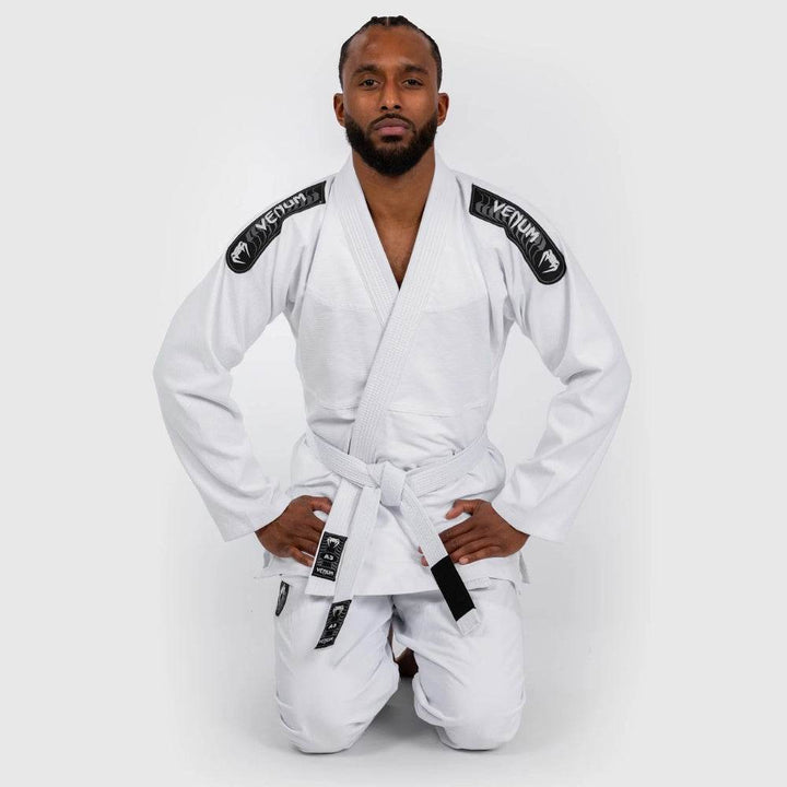 Martial artist kneels in a formal pose wearing the white Venum gi, displaying the gi's front fit and embroidered shoulder logos.