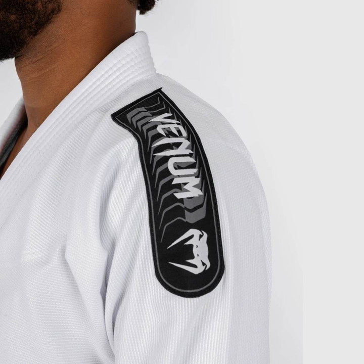 Detailed view of the Venum logo on the gi's shoulder, featuring black and silver embroidery on a white background with herringbone texture.