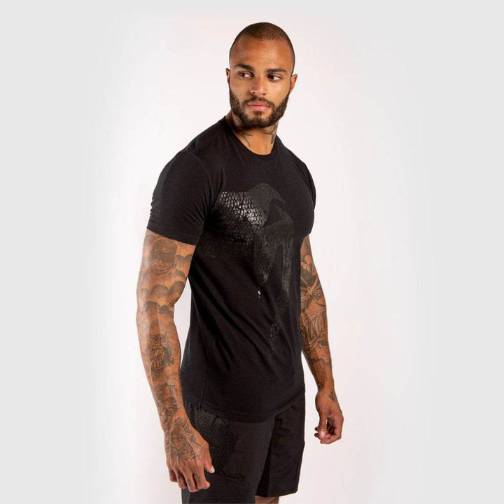 Side profile view of man in black snake-textured Venum t-shirt, standing with right arm extended back slightly.