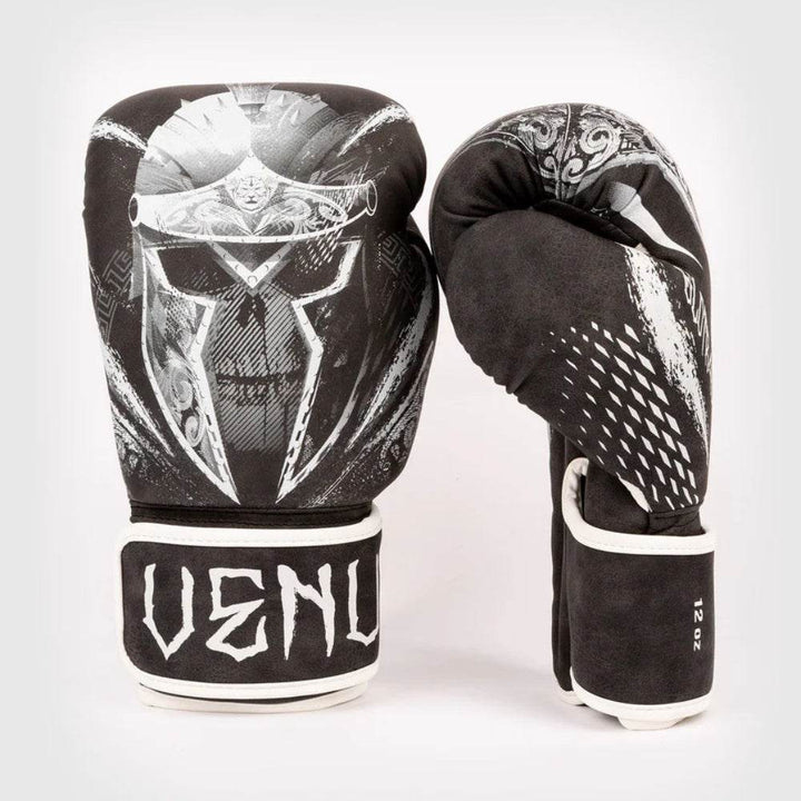 Front-and-side display of Venum Gladiator 4.0 boxing gloves, matte black with bold white Spartan-skull graphic and rough-edge “VENUM” wrist logo.