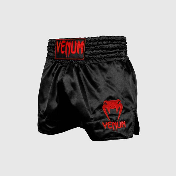 Black satin Muay Thai shorts with red embroidered Venum logo on left leg and waistband.