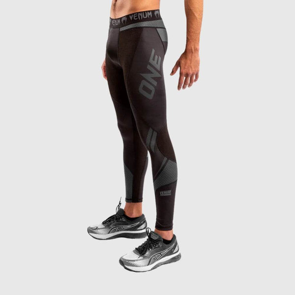 Male model wearing black compression spats with “ONE” branding in grey on right thigh, side mesh panels visible, paired with silver and black running shoes.