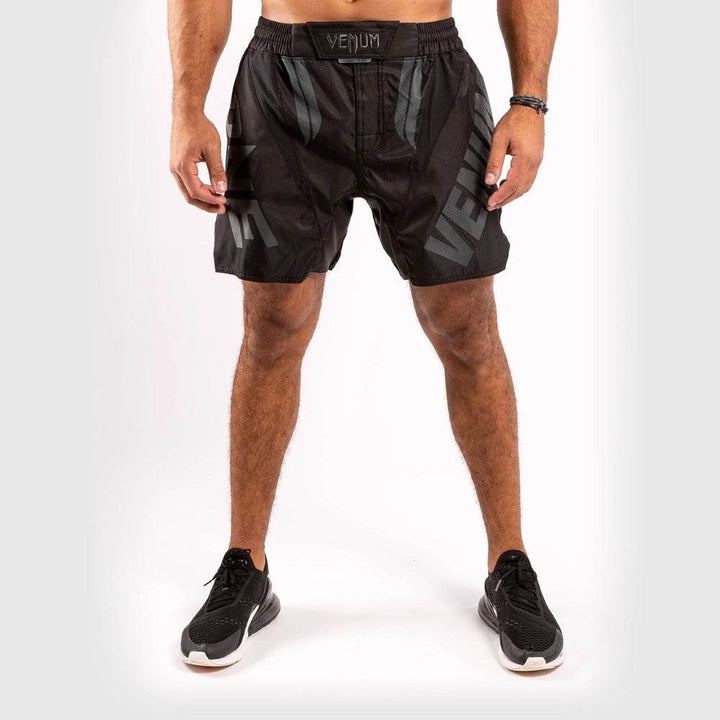 Man wearing Venum black fight shorts with subtle black "VENUM" and "ONE" branding on each leg, elastic waistband with Venum logo, standing front-facing.