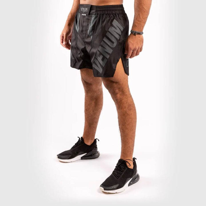 Angled front view of Venum black fight shorts showing the "VENUM" branding on the right leg, and side slit detailing, worn by model in black trainers.