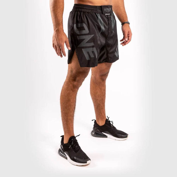 Angled front view highlighting the "ONE" branding on the left leg of black Venum fight shorts with textured gloss finish and side slit for mobility.