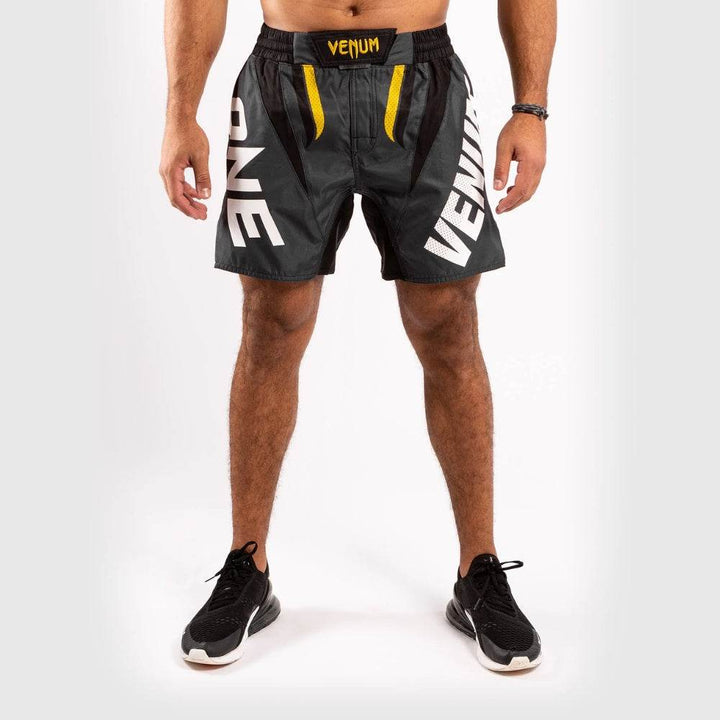 Black Venum fight shorts with “VENUM” and “ONE” text printed in large white letters on each leg. Yellow accents highlight the front waist area.