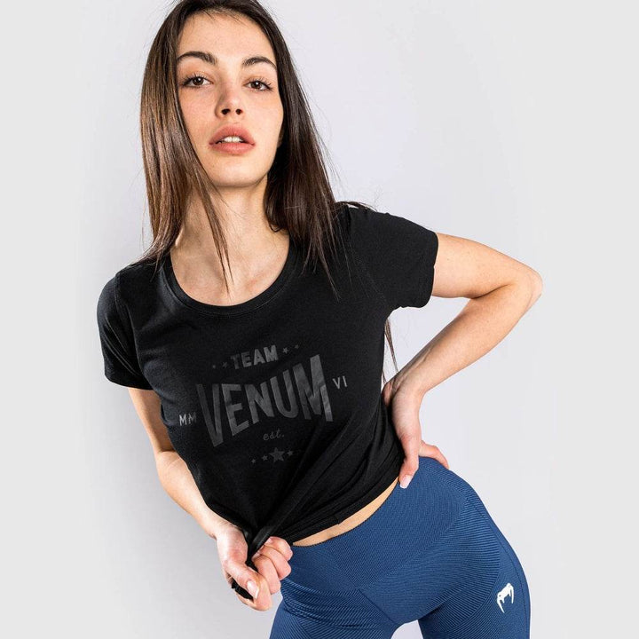 Woman wearing a black Venum Team t-shirt and blue leggings, slightly lifting the shirt to show fit.