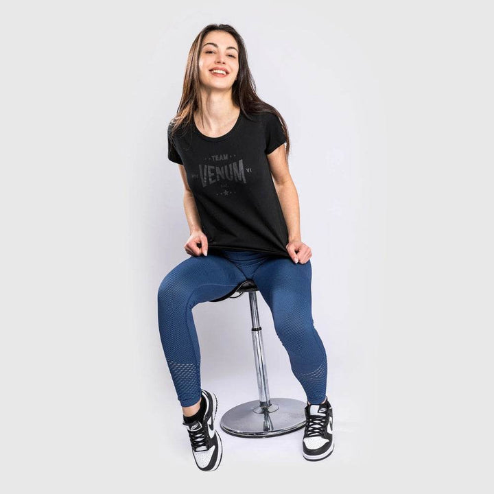 Woman sitting on a chrome stool in black Venum t-shirt and blue leggings, smiling casually.