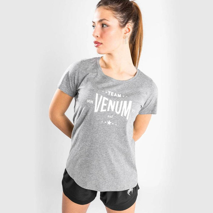 Woman in grey Venum Team t-shirt and black shorts, hands behind her back, looking sideways.