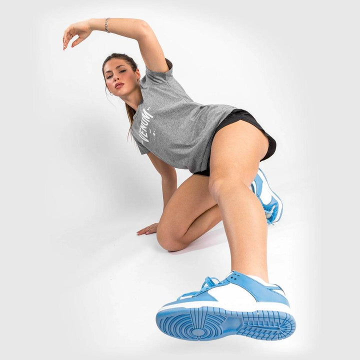 Woman in grey Venum Team t-shirt stretching backward from a floor pose, wearing white and blue sneakers.