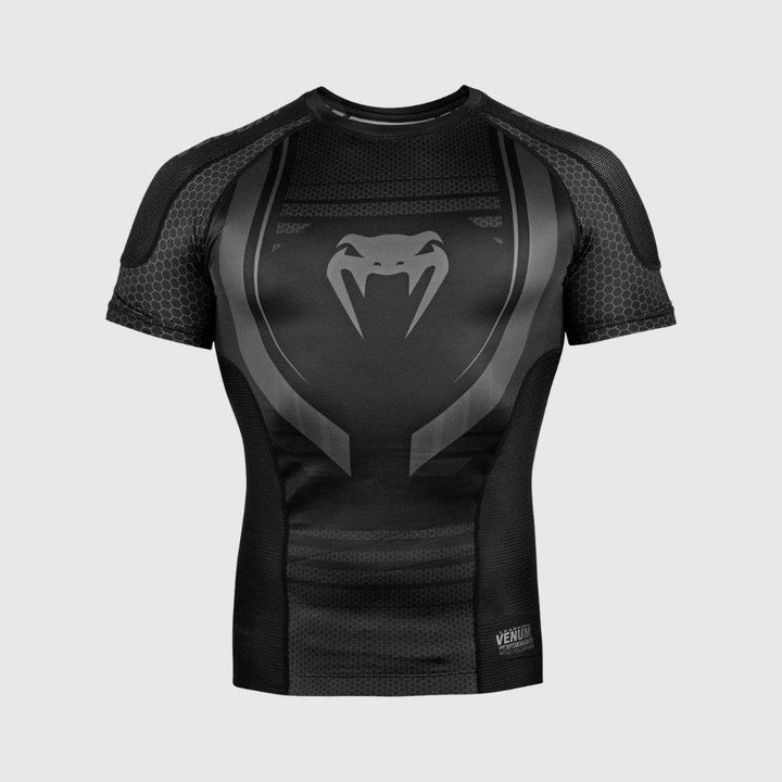 A black Venum short sleeve compression rash guard displayed against a light grey background, featuring a large grey Venum snakehead logo on the chest and a geometric pattern throughout the torso and sleeves.